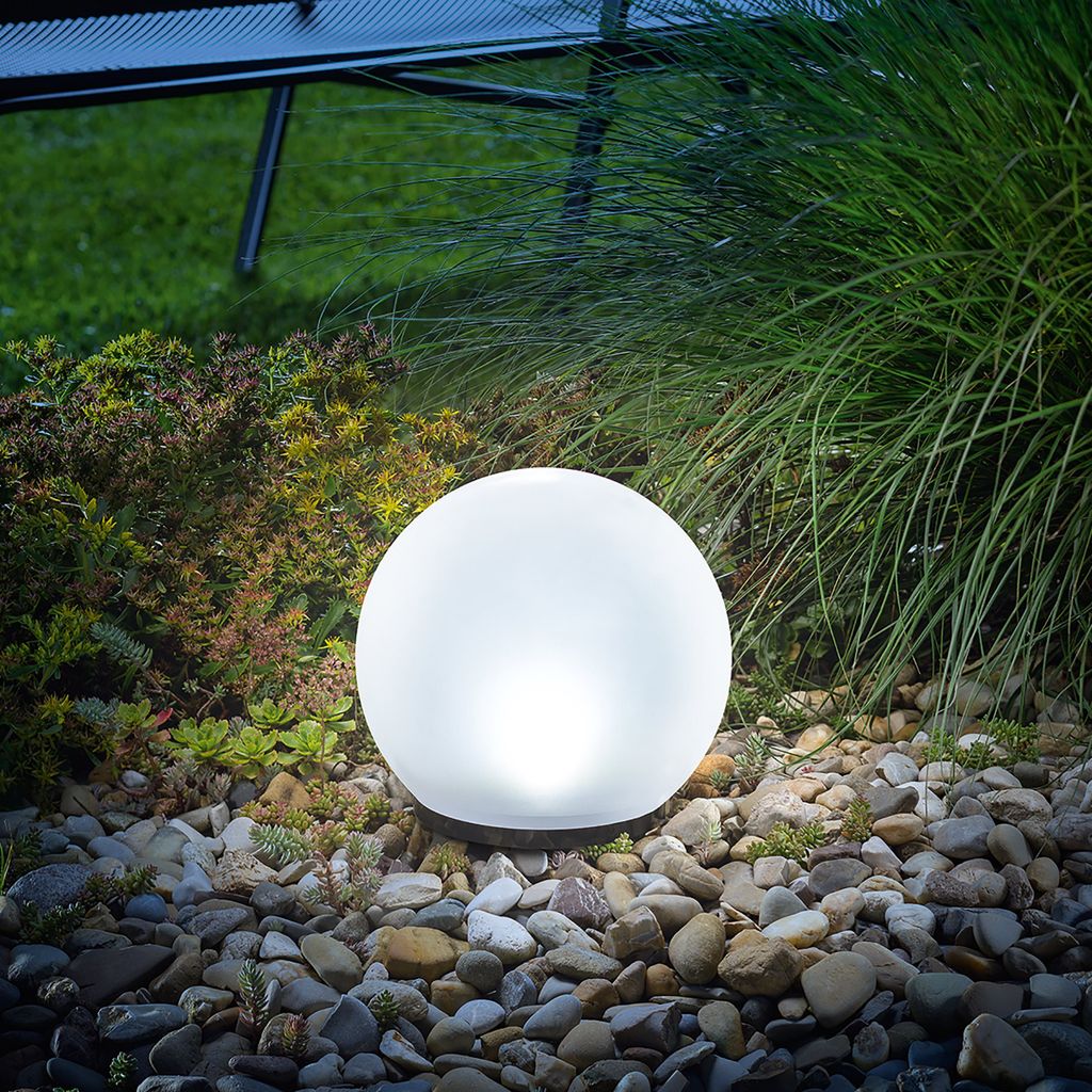 A glowing solar ball light sits in a garden bed surrounded by rocks and plants.