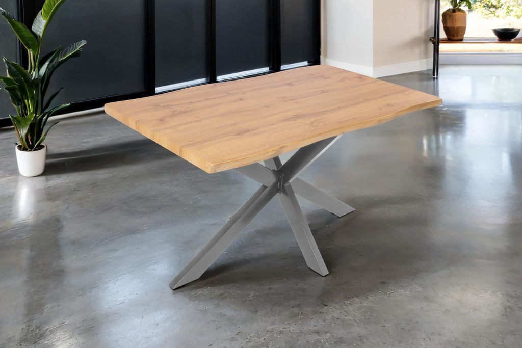 A wooden dining table with a metal spider base is shown in a room with a concrete floor and a dark wall.