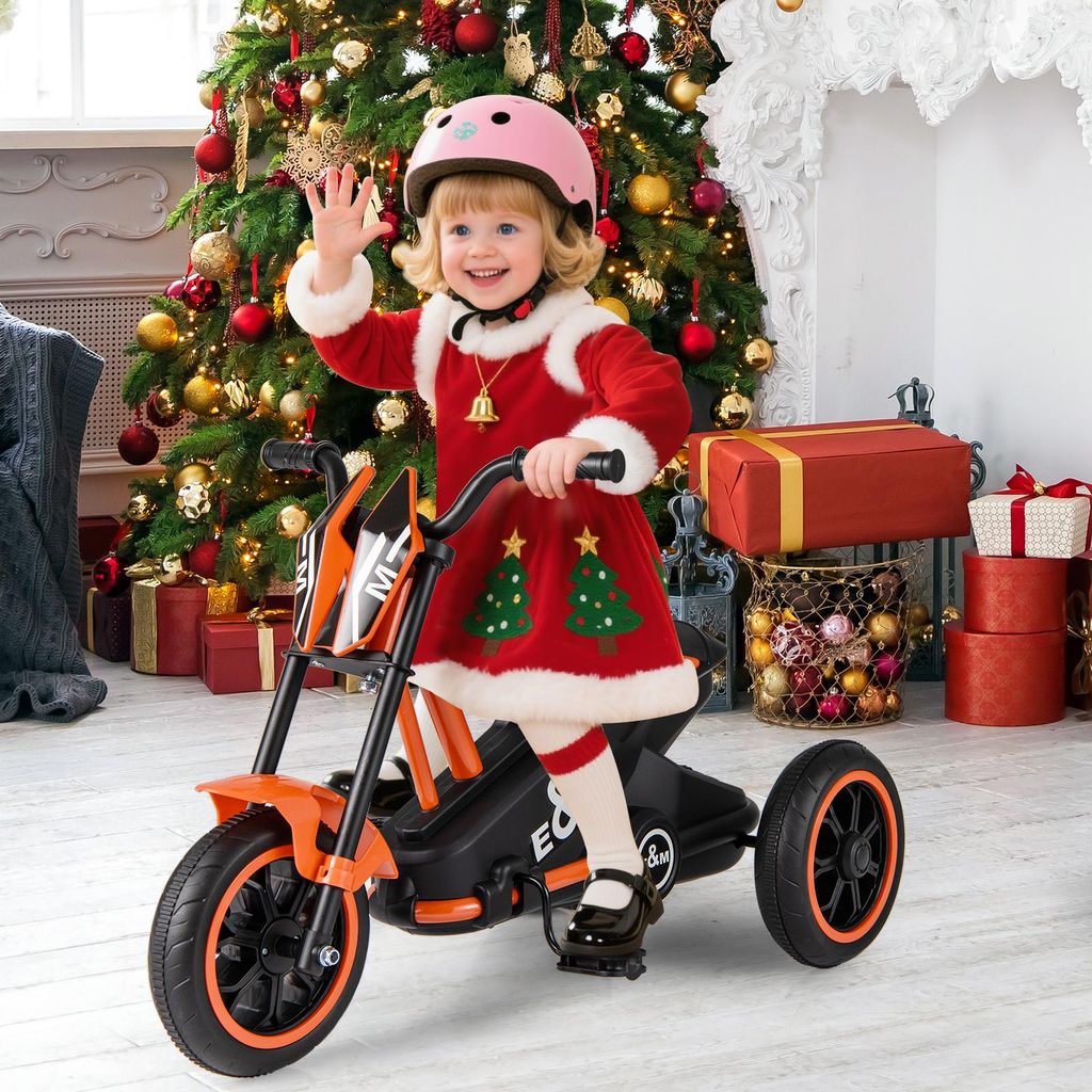 A young child dressed in a Santa-themed outfit rides an orange electric tricycle in front of a decorated Christmas tree and presents.