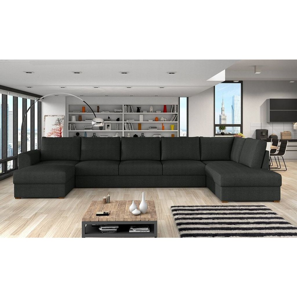 A large, dark gray sectional sofa is shown in a modern living room setting with a bookshelf and large windows in the background.
