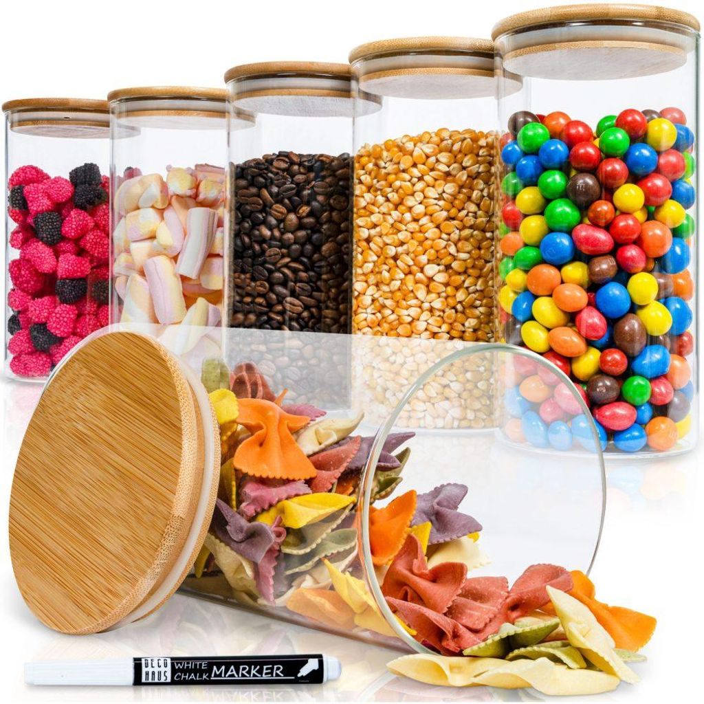Set aus sechs klaren Glas-Vorratsgläsern mit natürlichen Bambusdeckeln, gefüllt mit verschiedenen bunten Lebensmitteln, ein Glas umgekippt mit herausfallenden Farfalle-Nudeln, davor ein weißer Kreidestift