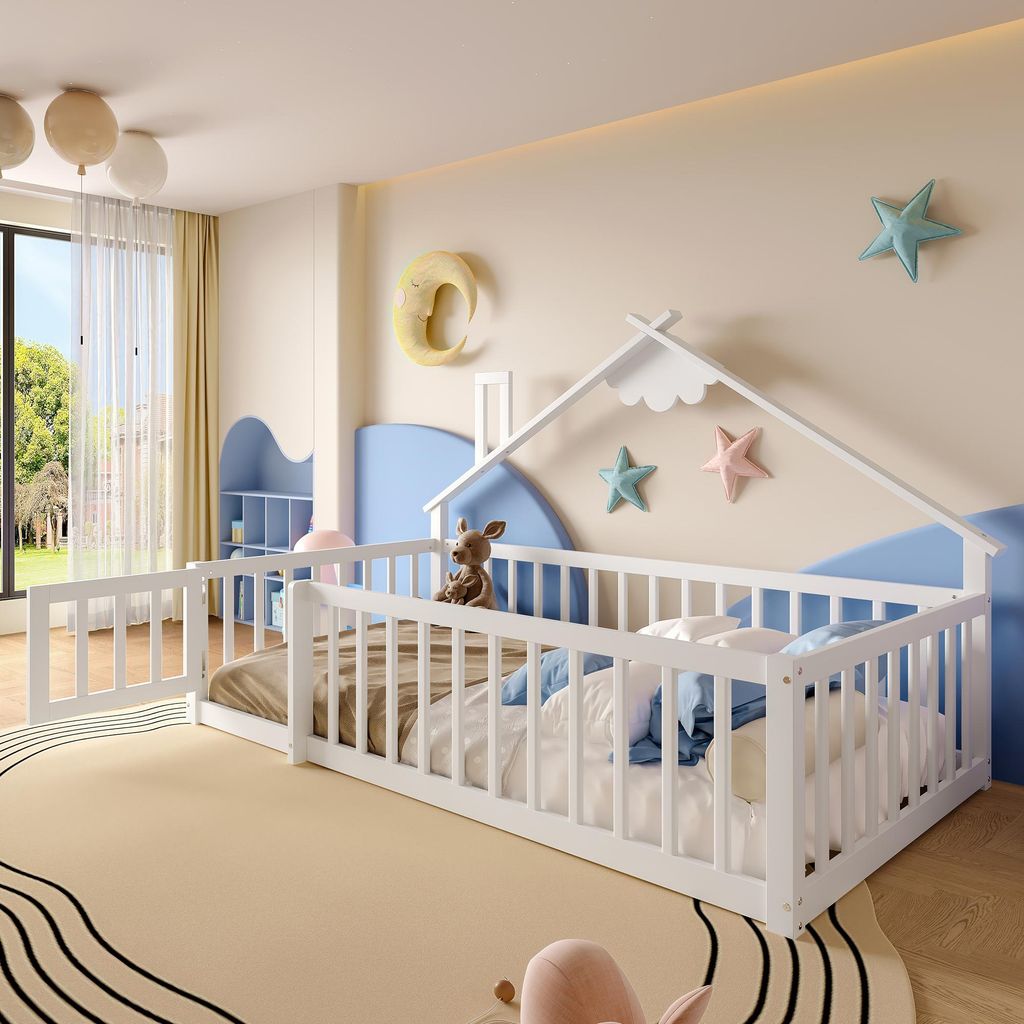A child's bedroom with a white wooden bed frame designed like a house, complete with a railing and a small fence. Toys and bedding are visible on the bed. The room is decorated with stars and a crescent moon on the wall, and a large window overlooks a gree