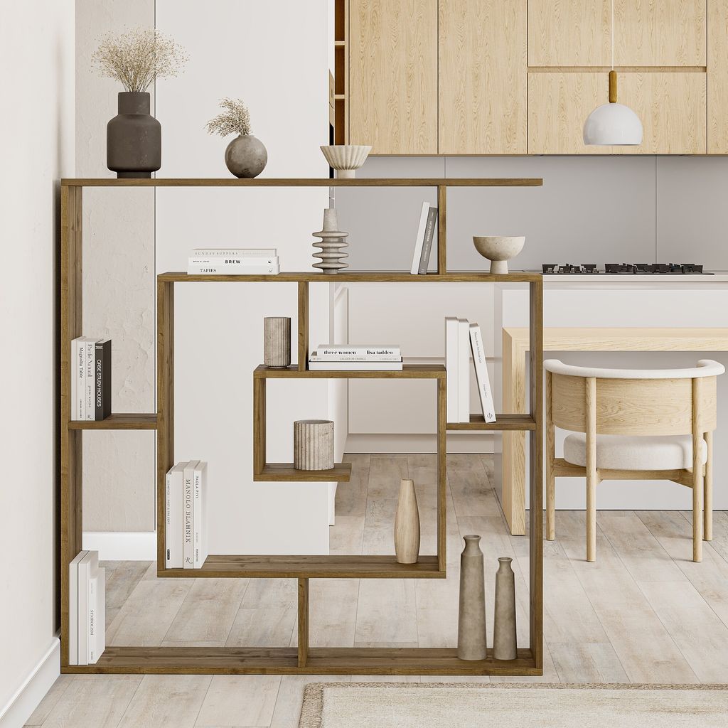A modern living room or dining area with a wooden bookshelf as a room divider, decorated with books and vases. A kitchen counter and stool are visible in the background.