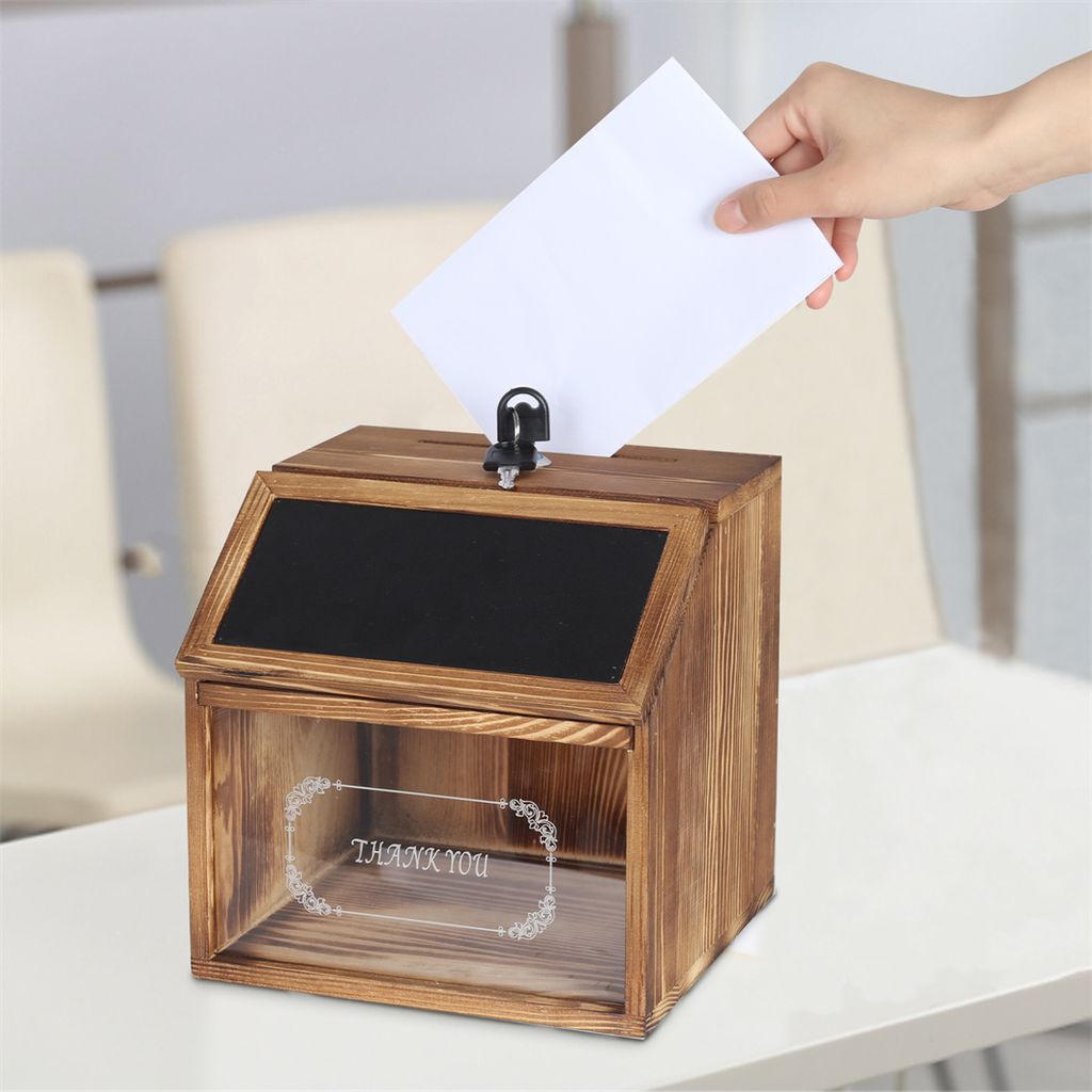 A hand is placing a white paper into a wooden suggestion box with a chalkboard top.