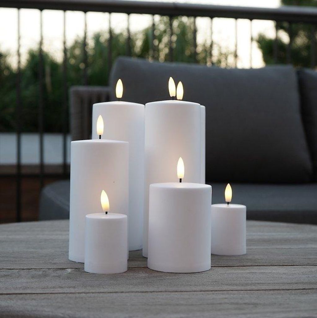 Several white LED candles of varying sizes are arranged on a wooden table outdoors, with a blurred background of outdoor furniture and greenery.
