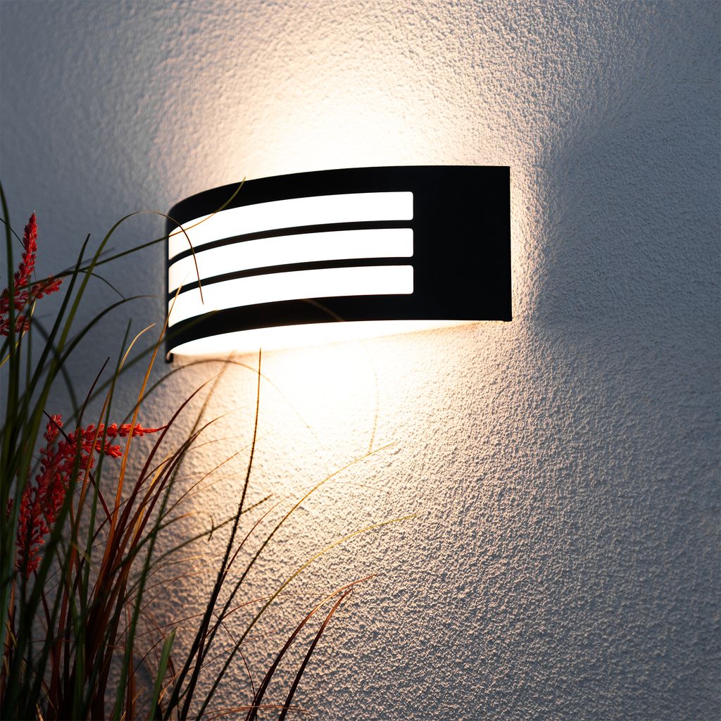 A black outdoor wall light with white illuminated strips is mounted on a textured white wall, with some tall grass and red flowers in the foreground.