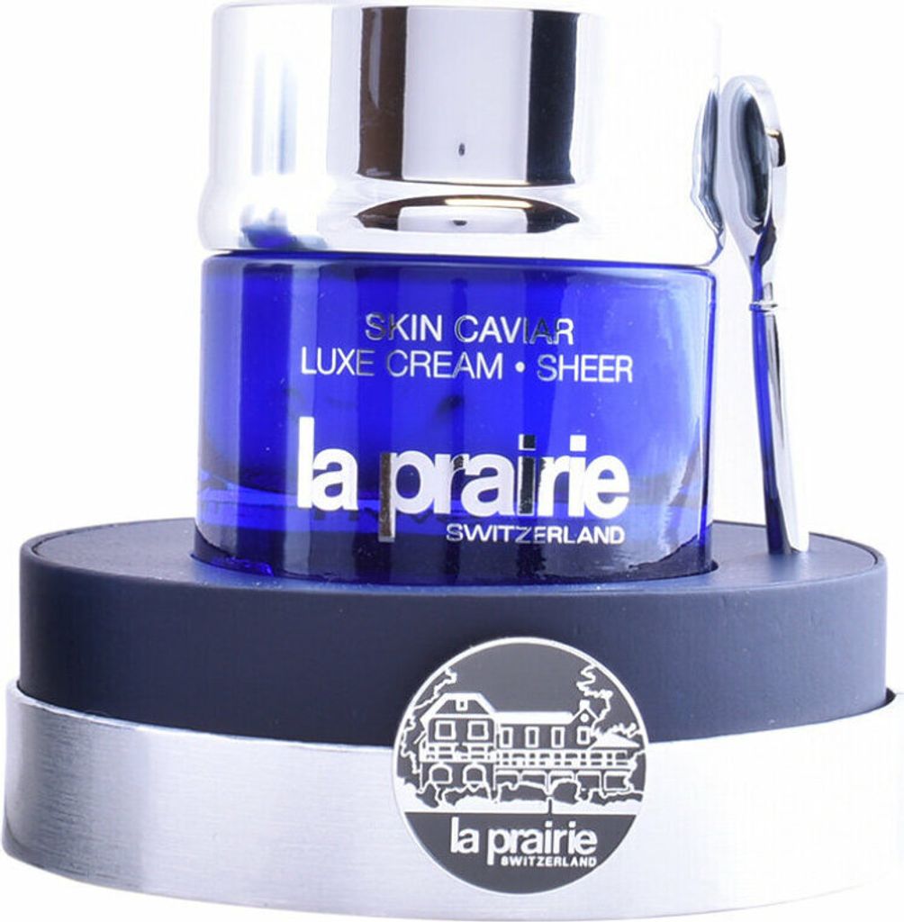 A jar of La Prairie Skin Caviar Luxe Cream Sheer is displayed on a dark blue and silver stand, with a silver spatula resting beside it. A circular emblem with a building illustration and the La Prairie logo is attached to the stand.