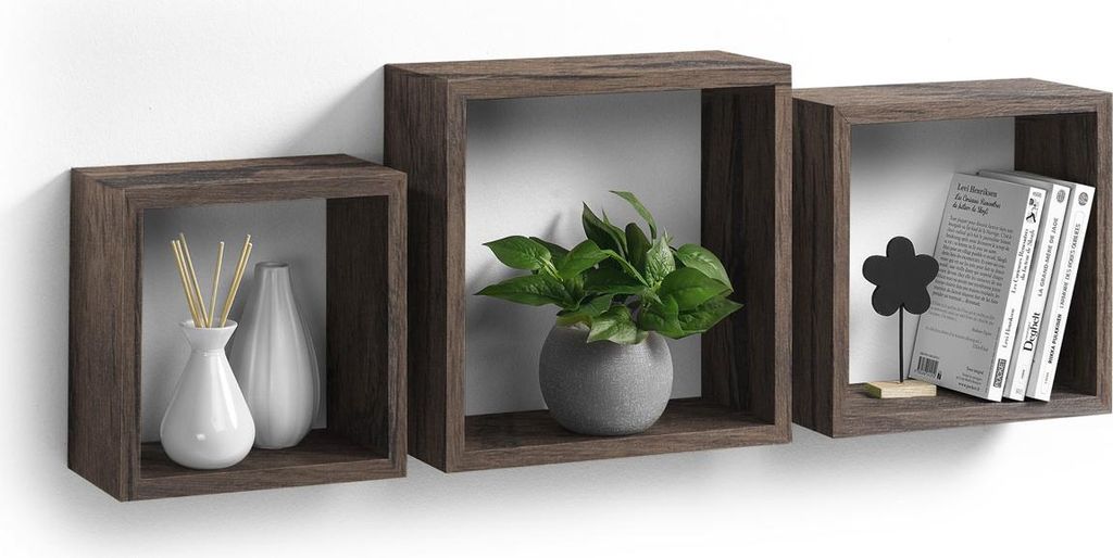 Three dark brown floating cube shelves are mounted on a white wall. The left shelf contains two white vases, one with reeds. The middle shelf holds a green plant in a gray pot. The right shelf displays a black flower decoration and three books.