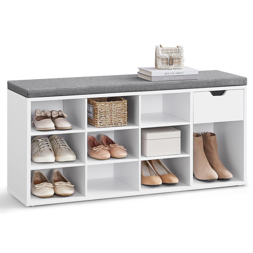 A white shoe bench with a gray upholstered top, featuring multiple compartments and a drawer, displaying various shoes and a decorative basket and box.