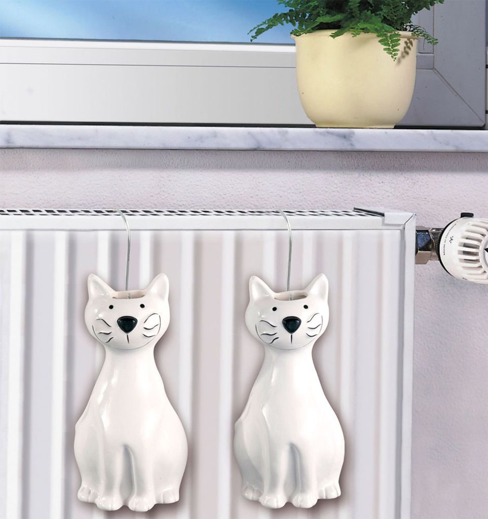 Two white ceramic cat-shaped humidifiers hang on a radiator, with a potted plant visible on a windowsill in the background.
