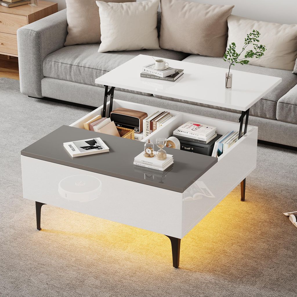 A modern living room scene featuring a multifunctional coffee table with its top lifted, revealing storage compartments. The table is placed in front of a sofa, with decorative items and books on display.