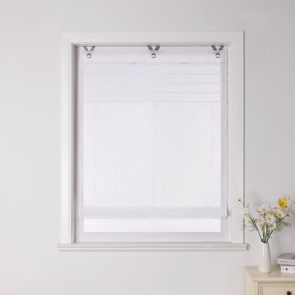A white Roman blind is installed in a white window frame against a white wall. A small vase with flowers sits on a bedside table to the right of the window.