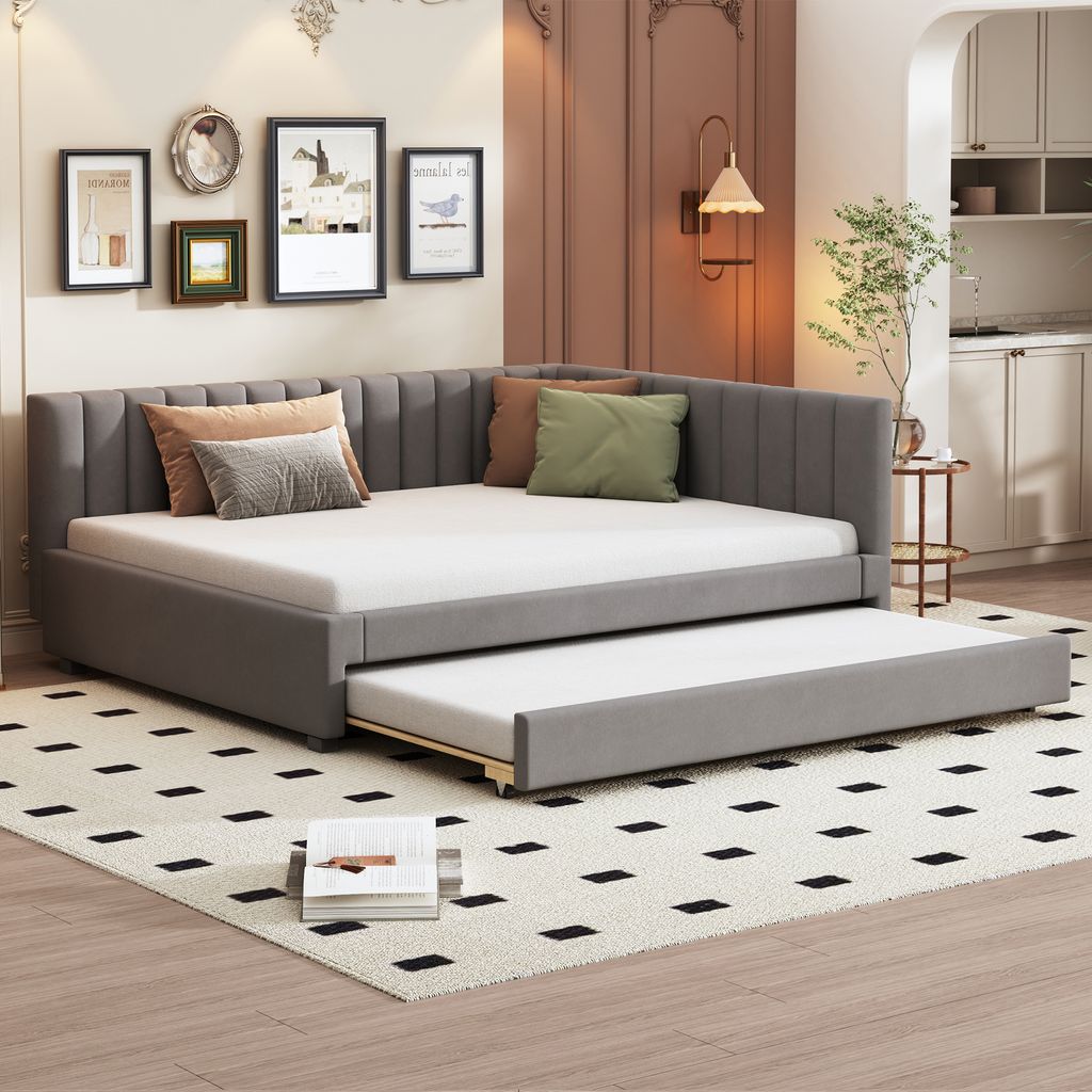 A modern living room with a grey upholstered daybed with a pull-out trundle bed, decorated with throw pillows. The room also features framed artwork on the wall and a small side table with a lamp.