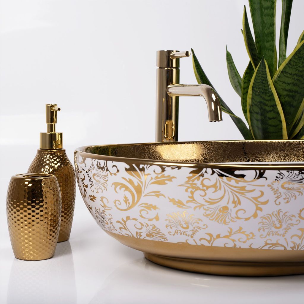 A close-up shot of a decorative washbasin with gold accents and floral patterns, accompanied by a gold soap dispenser and a toothbrush holder. A snake plant is visible in the background.