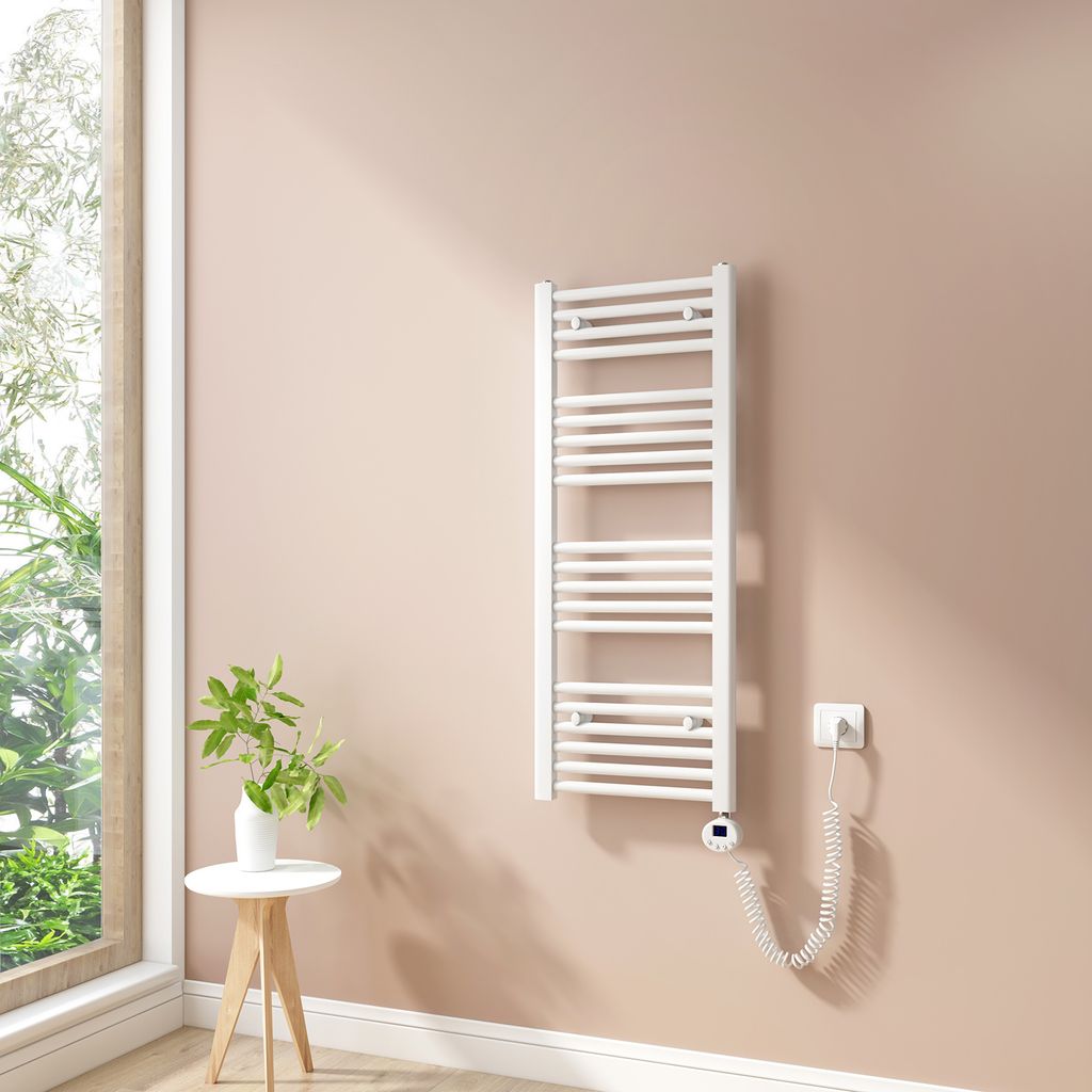A white electric towel radiator mounted on a light pink wall next to a large window. A small white side table with a green plant in a white vase is to the left of the radiator.