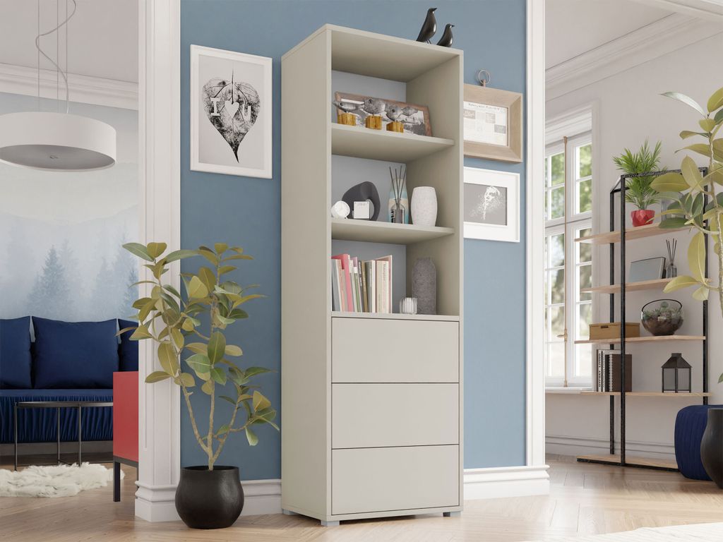 A tall, narrow bookshelf with three drawers is shown in a well-lit room with blue walls. A plant is in the foreground, and other furniture and decor are visible in the background.