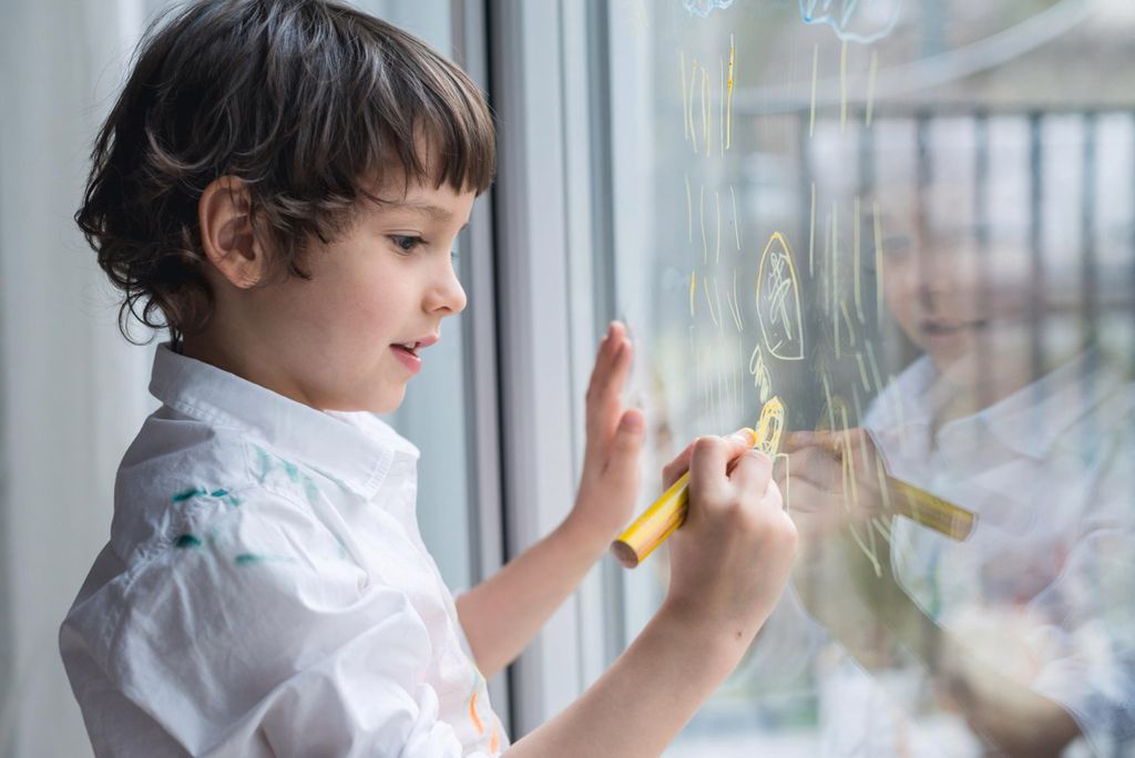 A young boy drawing on a window with a yellow crayon.