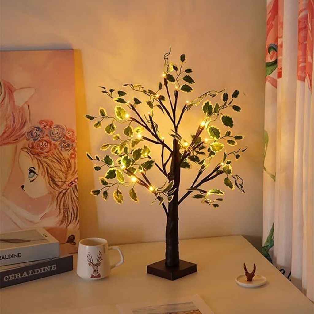 A decorative artificial tree with lights on a desk, next to a painting and a mug.
