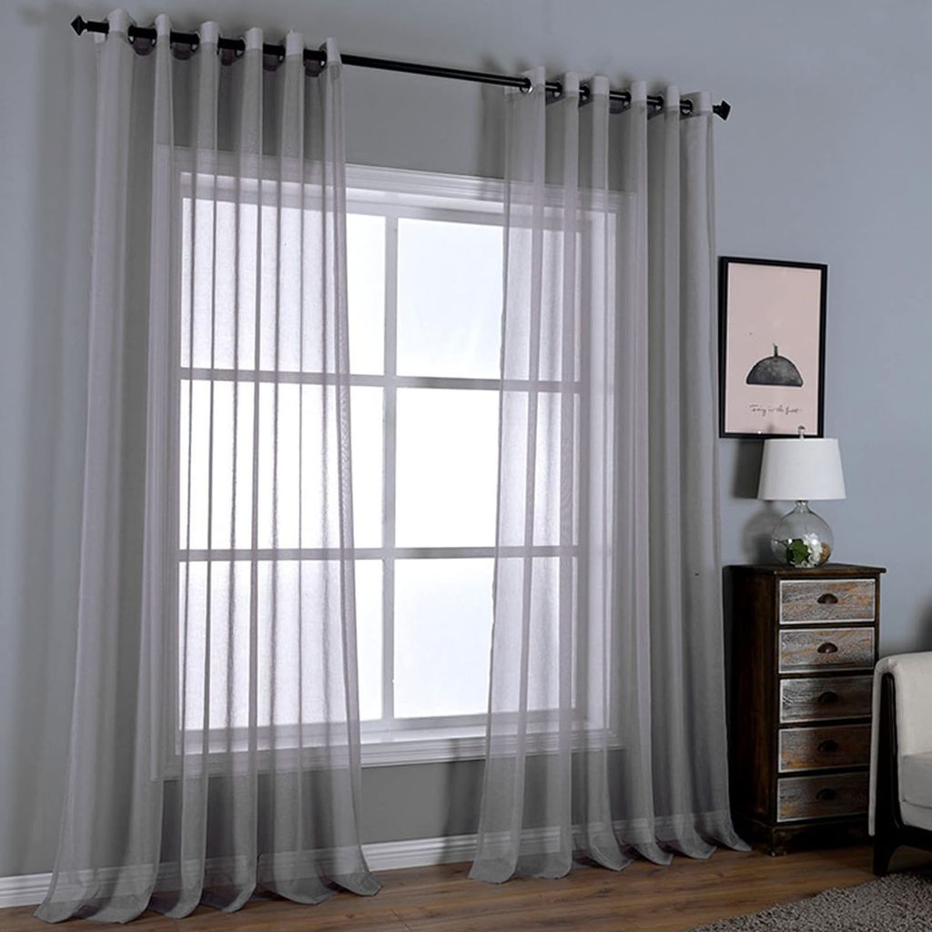 A window with light gray sheer curtains hanging in front of it, with a small wooden dresser and a lamp to the right.