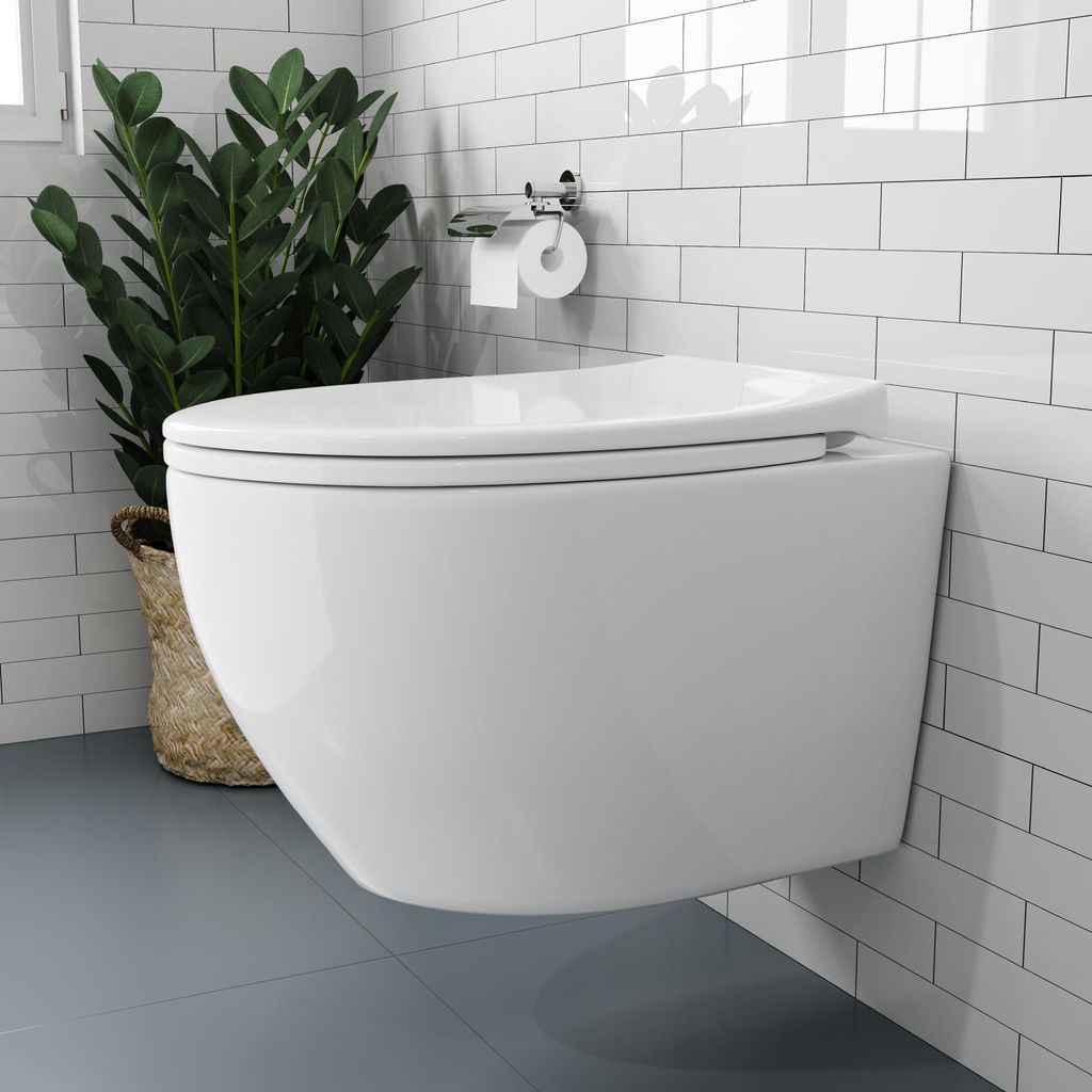 A modern white wall-mounted toilet in a bathroom with white subway tiles and gray flooring. A potted plant is visible to the left, and a toilet paper holder with a roll is mounted on the wall.