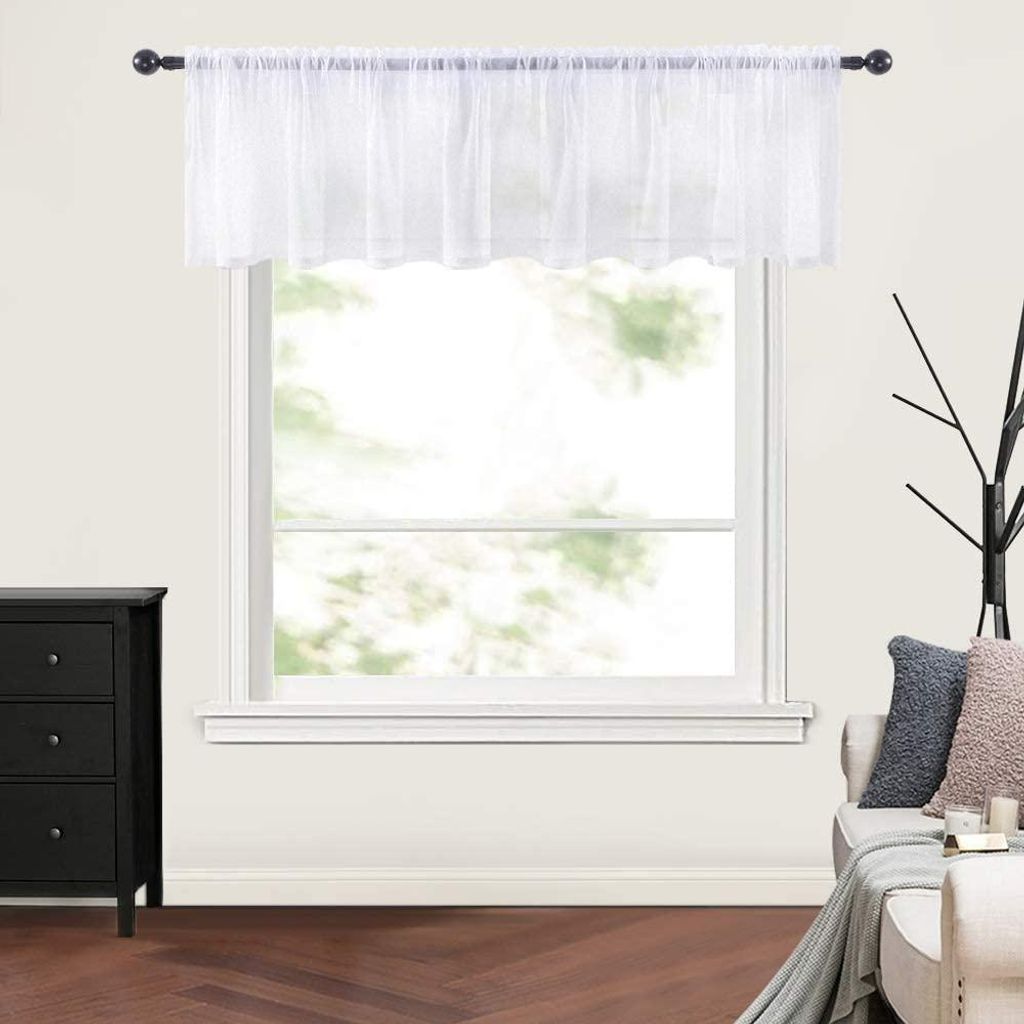 A white sheer valance is hung on a curtain rod above a window. The window looks out onto trees and greenery. To the left is a black dresser, and to the right is a sofa with pillows.