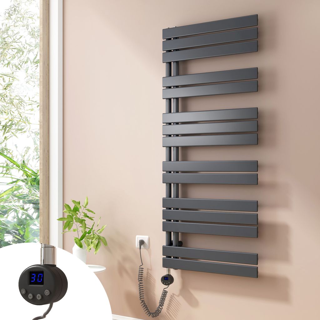 A dark gray electric towel radiator is mounted on a light pink wall next to a window with greenery visible outside. A small black digital thermostat with a blue display is attached to the radiator.