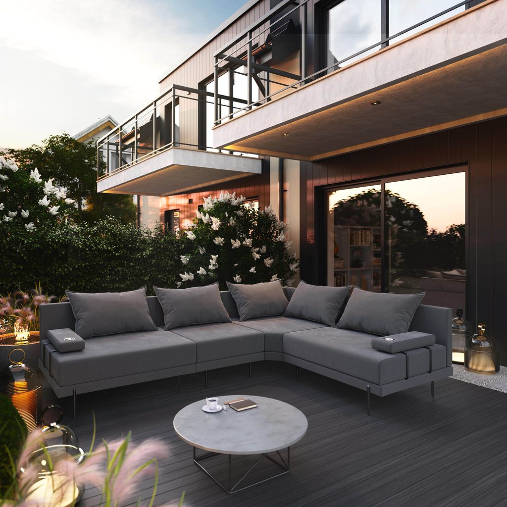 A modern outdoor lounge set on a dark wooden deck, with lush greenery and a contemporary building in the background during sunset.