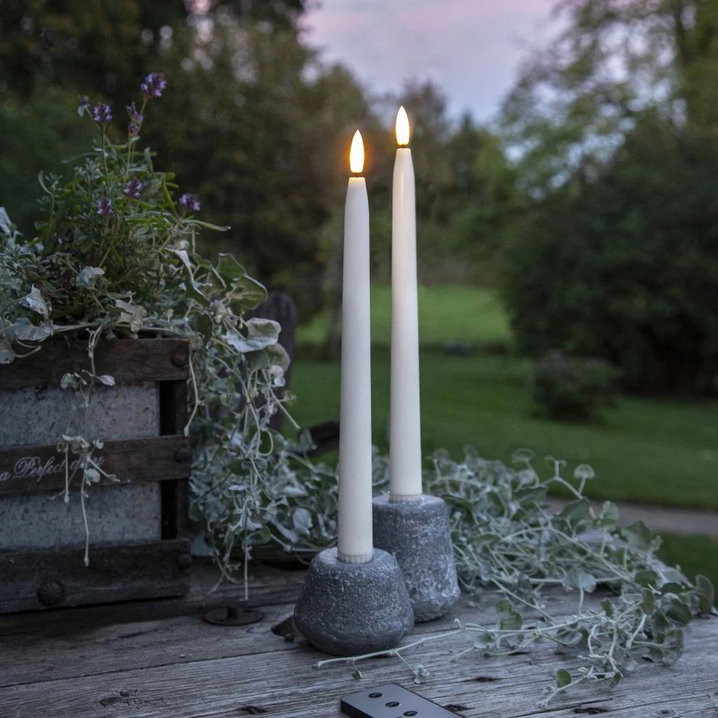 Two LED candles with flickering flames stand on a wooden table in an outdoor setting, with greenery and trees in the background.