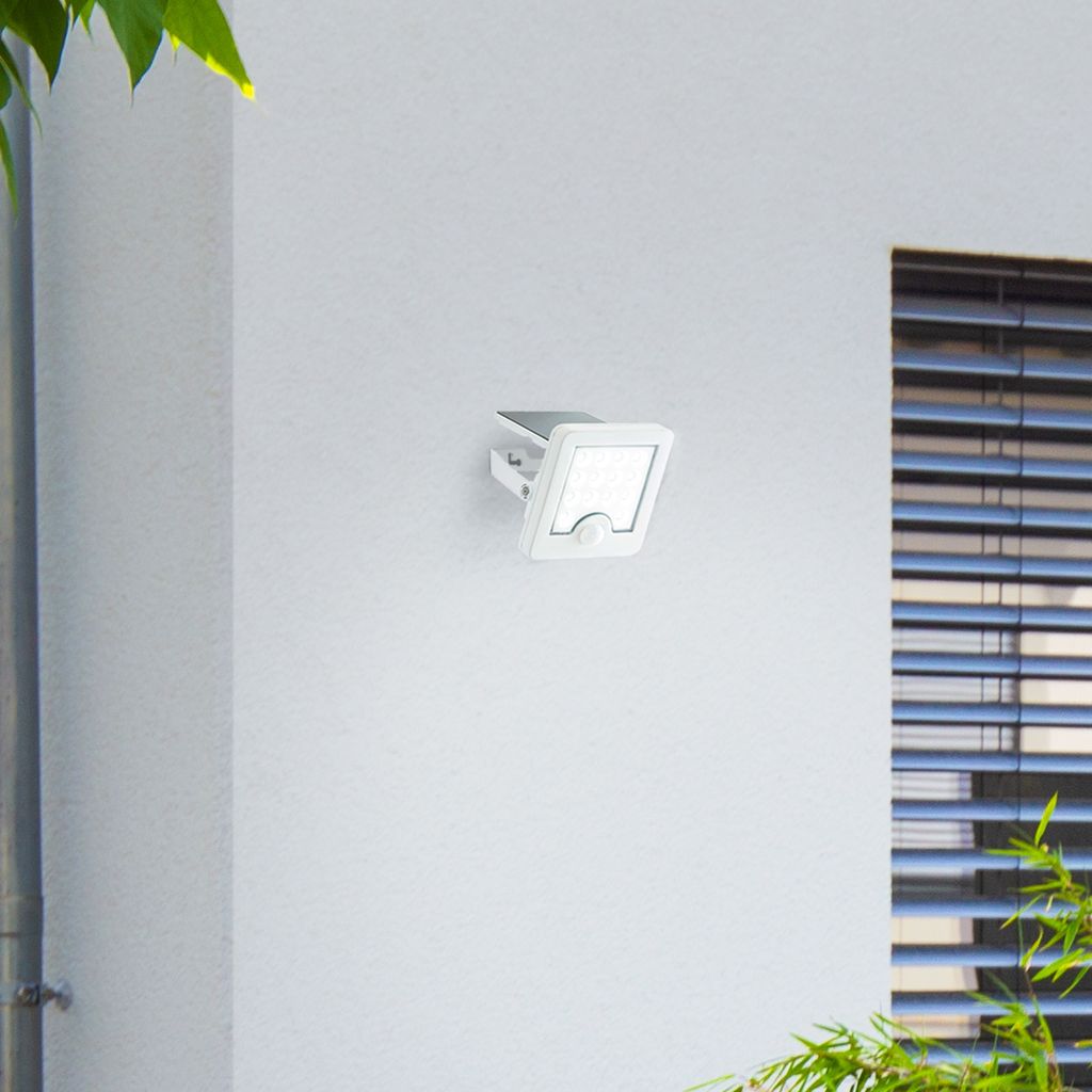 A white solar outdoor wall light with a motion sensor is mounted on a white exterior wall next to a window with blinds and some greenery.