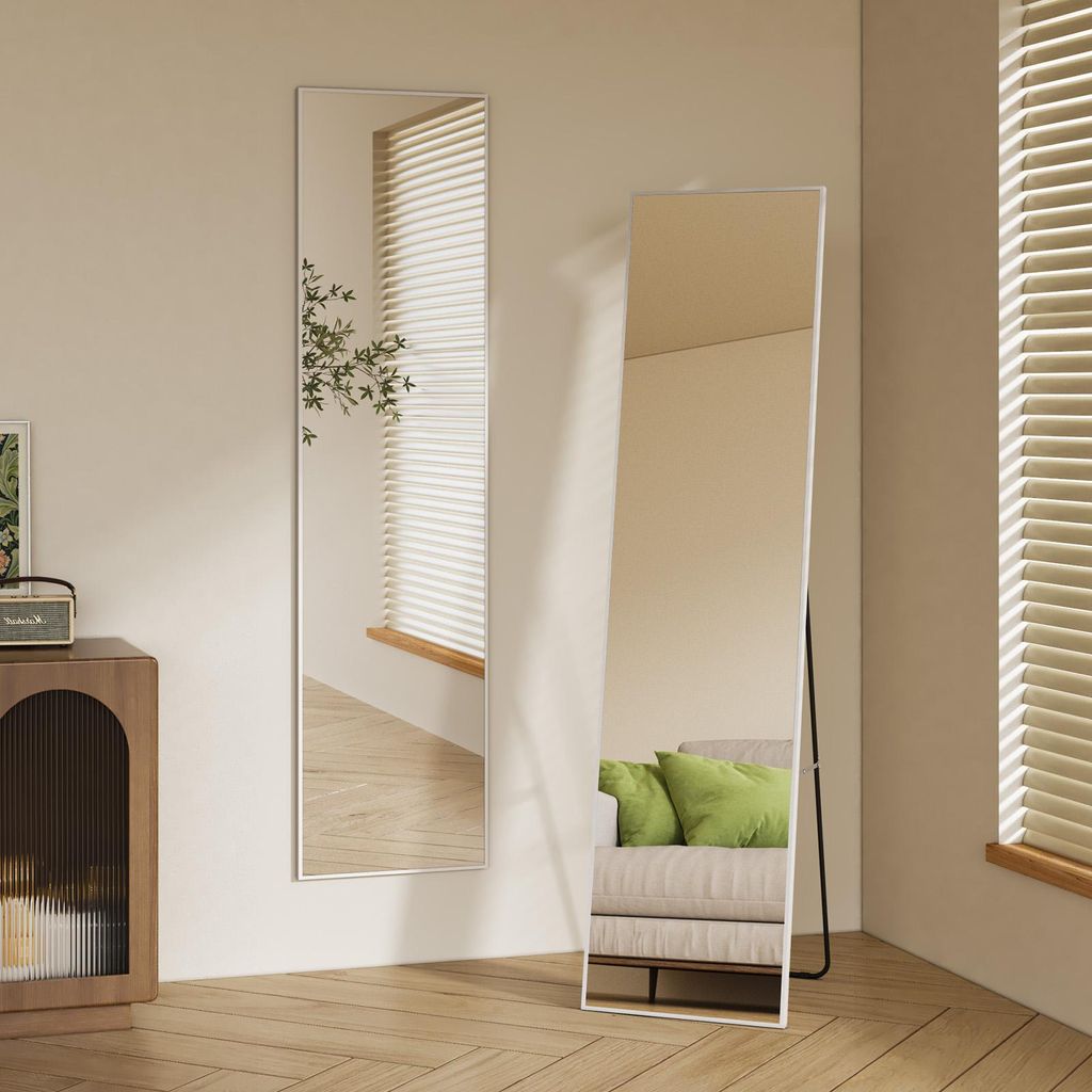 Two full-length mirrors are shown in a room with wooden floors and blinds on the windows. One mirror is leaning against the wall, and the other is standing on a black metal frame. A sofa with green cushions is reflected in the standing mirror.