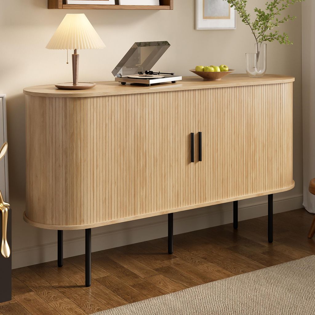 A modern wooden sideboard with black legs, featuring a ribbed texture and two black handles. It is placed in a room with a wooden floor and a beige rug. A lamp, a record player, and a bowl of fruit are on top of the sideboard.