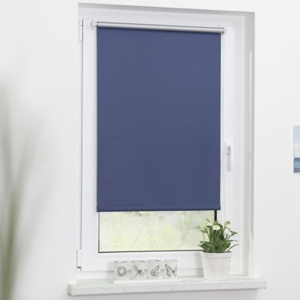 A blue roller blind is installed on a white window frame. A small potted plant with white flowers sits on the windowsill below the blind, along with three decorative glass candle holders.
