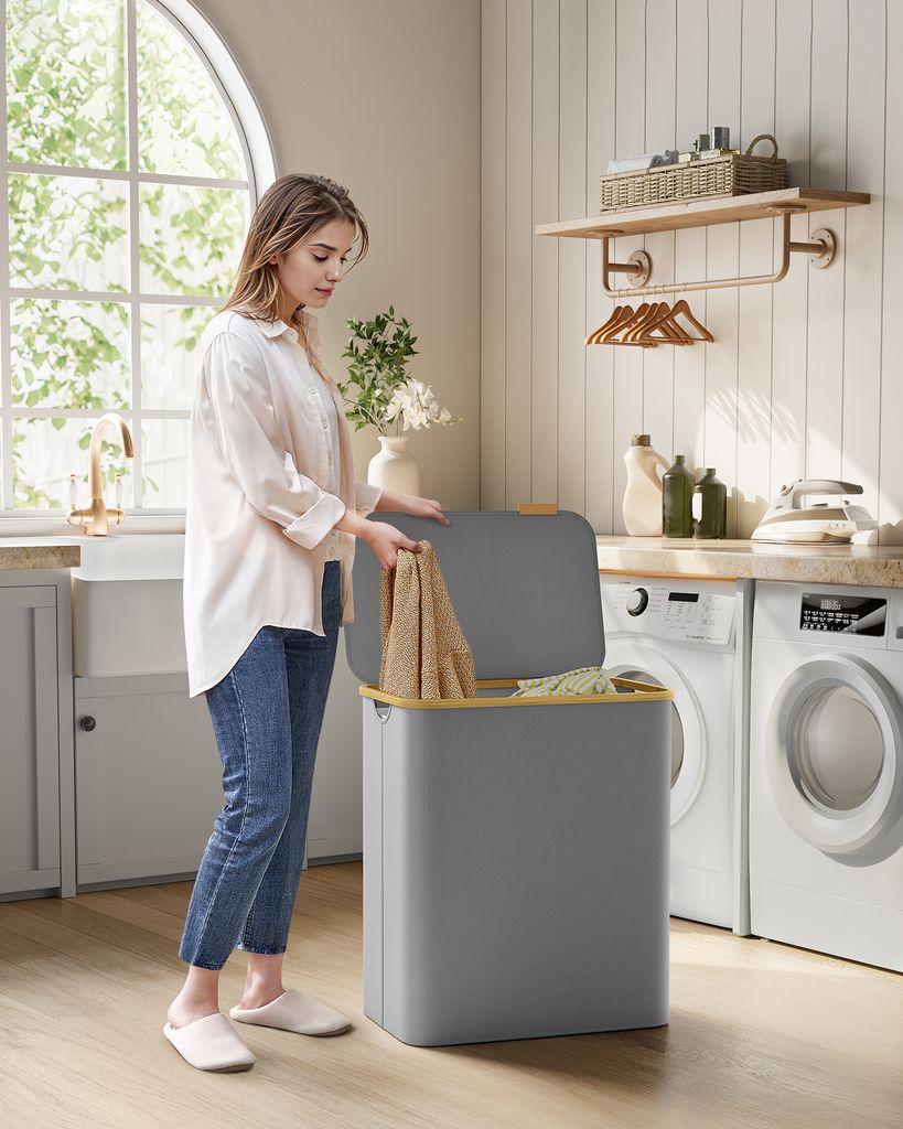 A woman is putting laundry into a gray laundry basket with a lid in a laundry room.