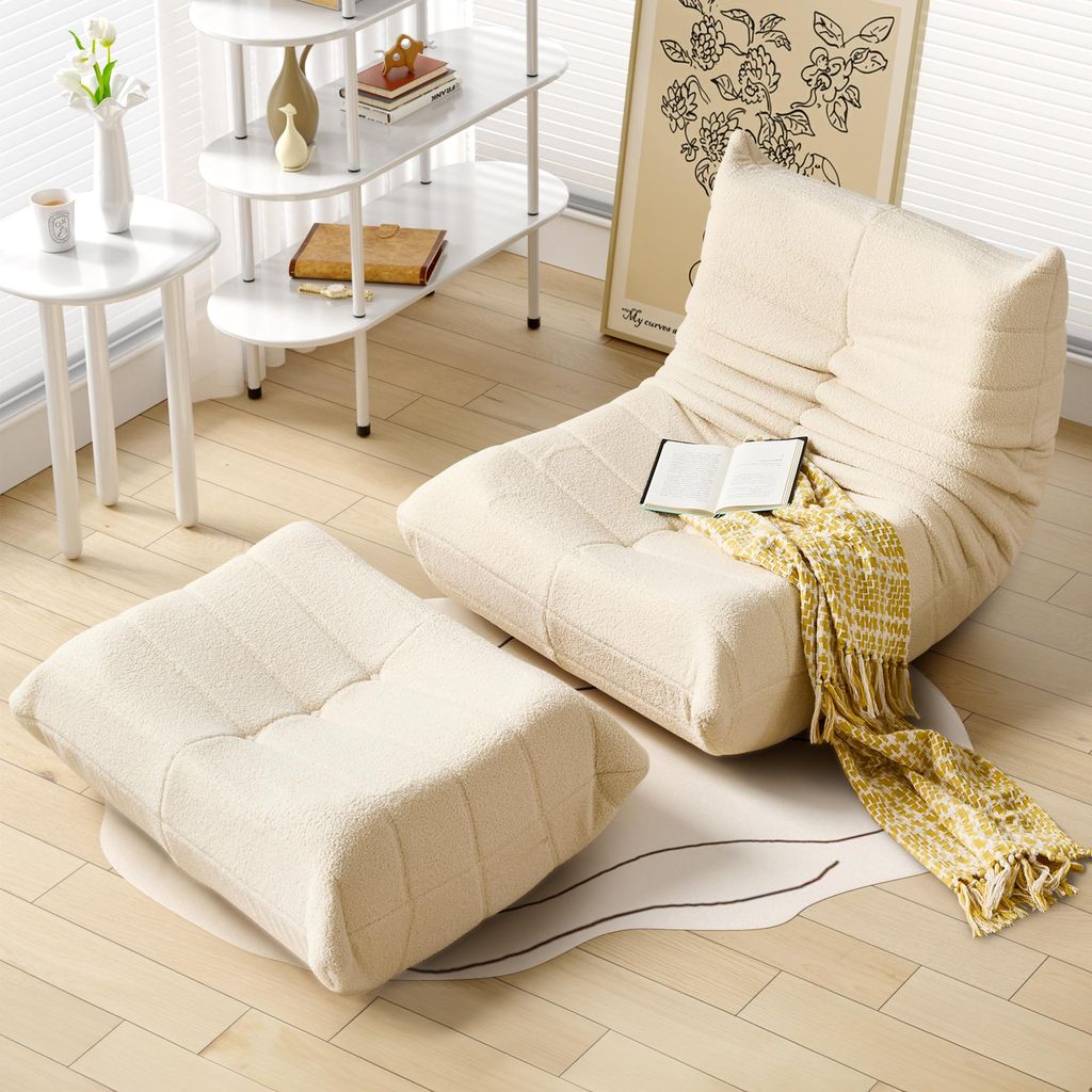 A beige sofa and ottoman set are arranged on a rug on a wooden floor, with a small side table and shelving unit in the background.