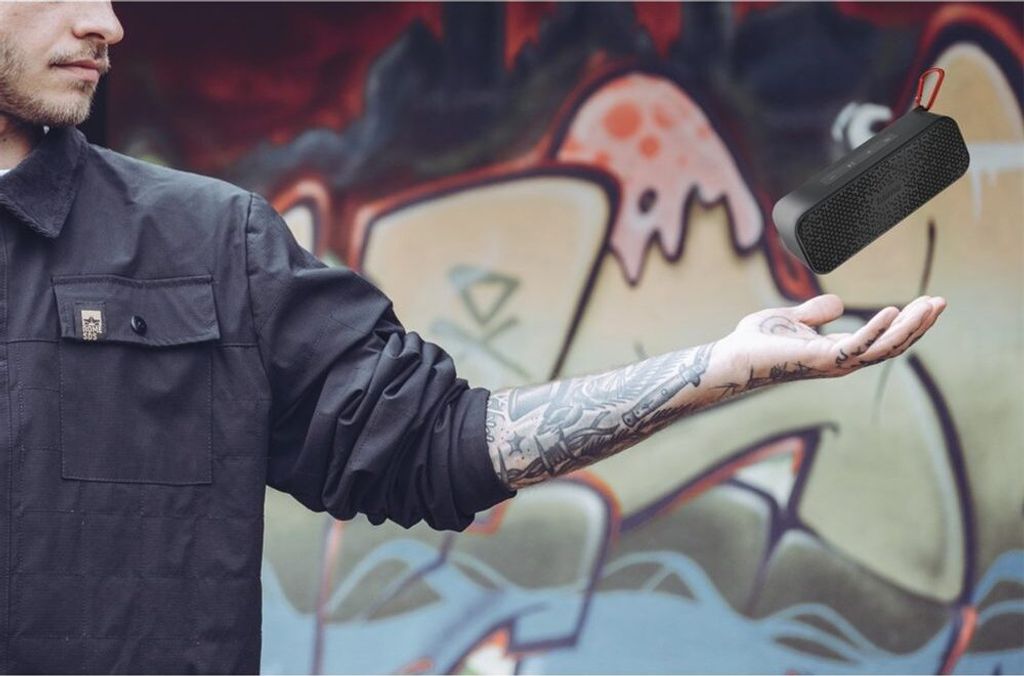 A person holding a Bluetooth speaker in front of a graffiti-covered wall.