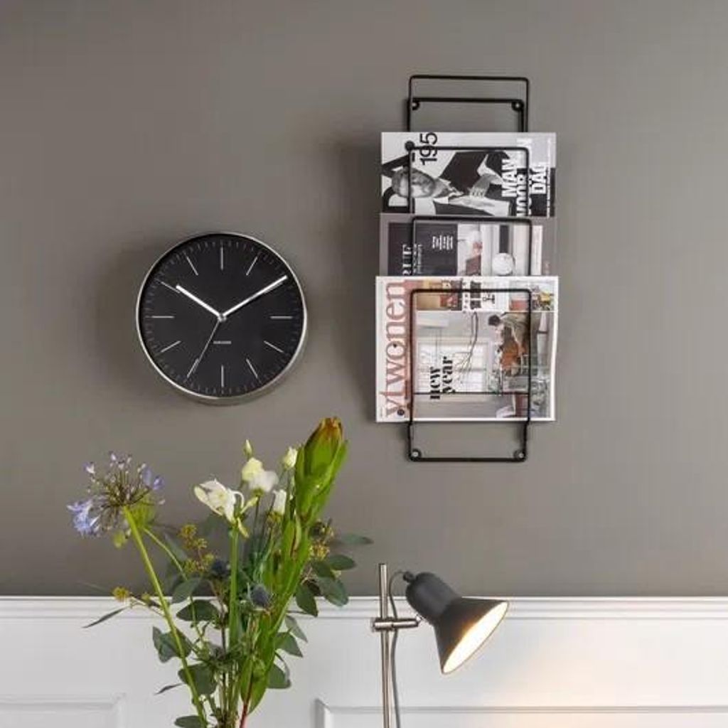 A wall with a clock and a magazine rack, with flowers and a lamp in the foreground.