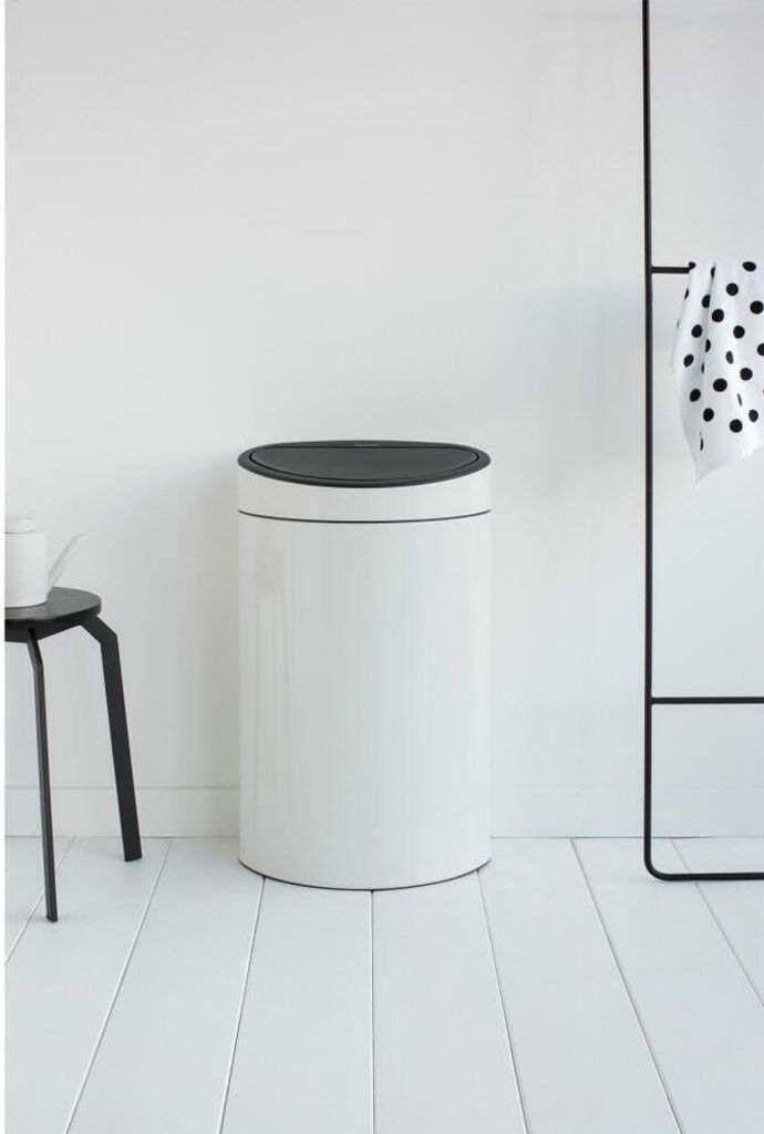A white Brabantia Touch Bin Mülleimer with a black lid stands on a white wooden floor against a white wall. A black stool with a white pitcher is to the left, and a black towel rack with a black and white polka dot towel is to the right.