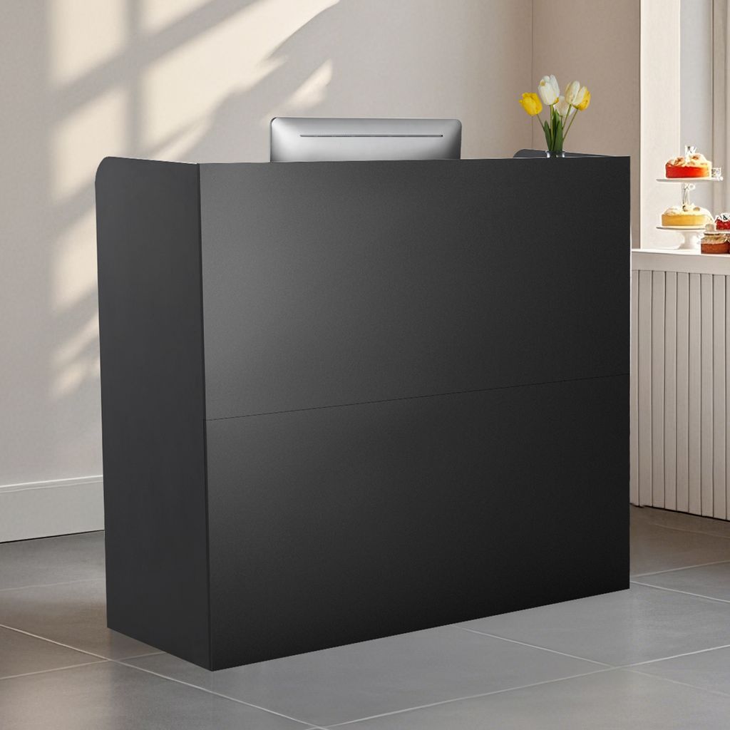 A black reception desk with a computer monitor on top, placed in a well-lit room with sunlight casting shadows on the wall.