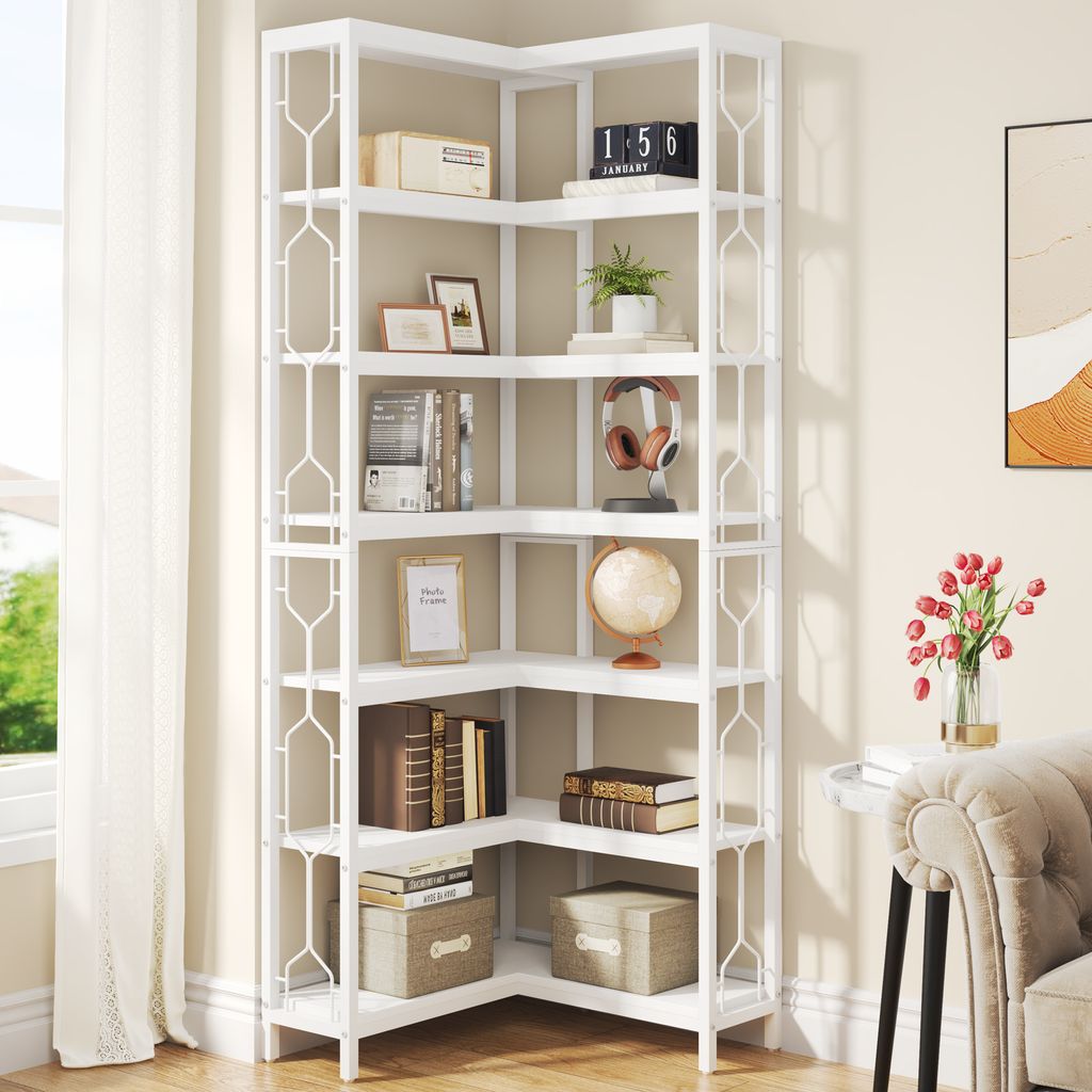 A white corner bookshelf filled with books and decorative items, next to a window with curtains and a part of a sofa and flowers in a vase.