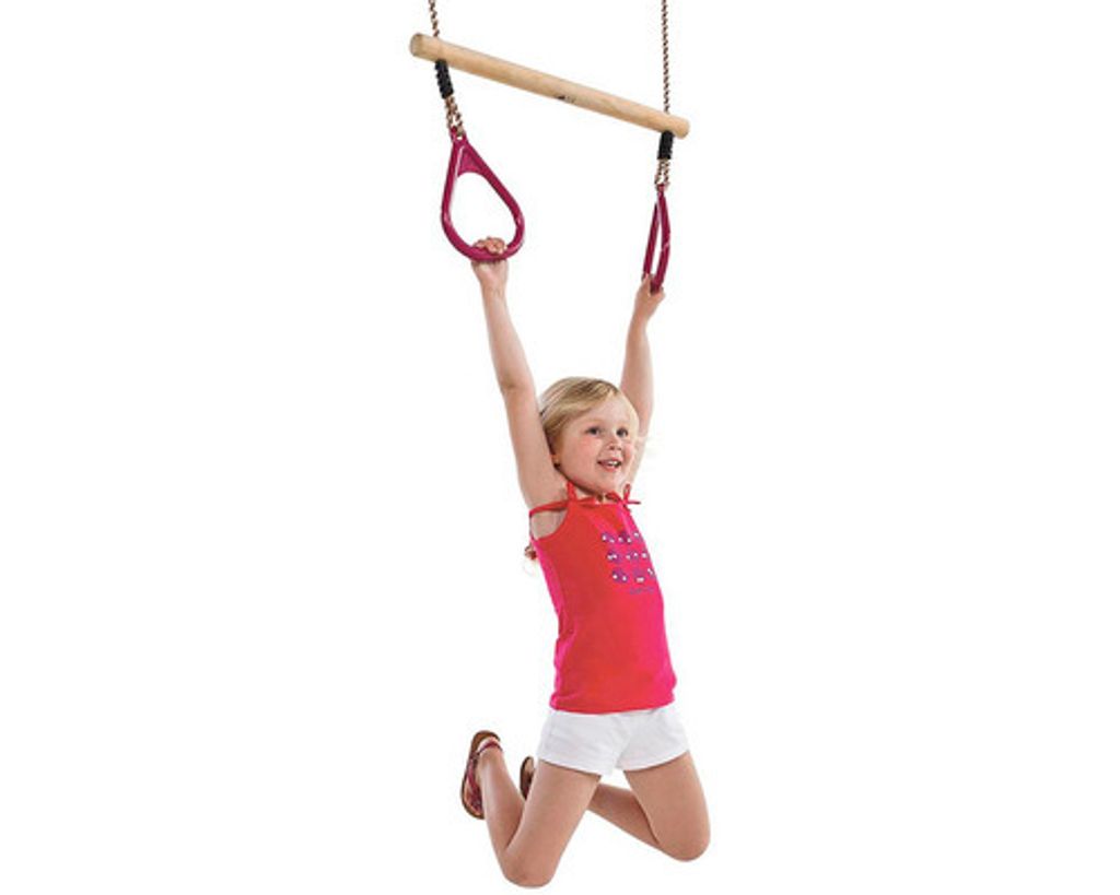 A young girl is hanging from a wooden trapeze bar with red rings.