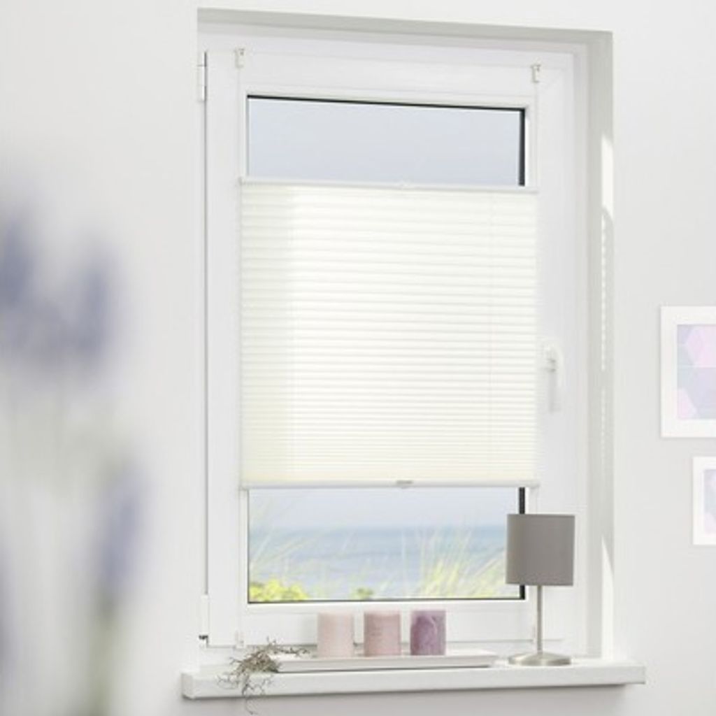 A white pleated blind is installed on a window overlooking a beach scene. There are three candles and a small lamp on the windowsill.