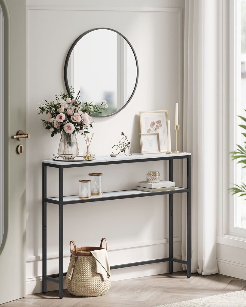 A narrow console table with two shelves, decorated with flowers, picture frames, and a basket, placed in a hallway next to a door and window.