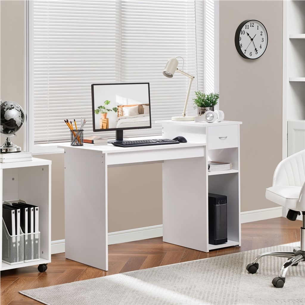 A white computer desk with a monitor, keyboard, lamp, and storage shelves is set up in a room with blinds and a clock on the wall.
