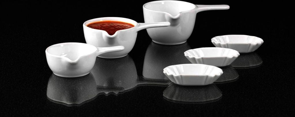 A set of white ceramic dipping bowls and sauce boats are arranged on a reflective black surface.