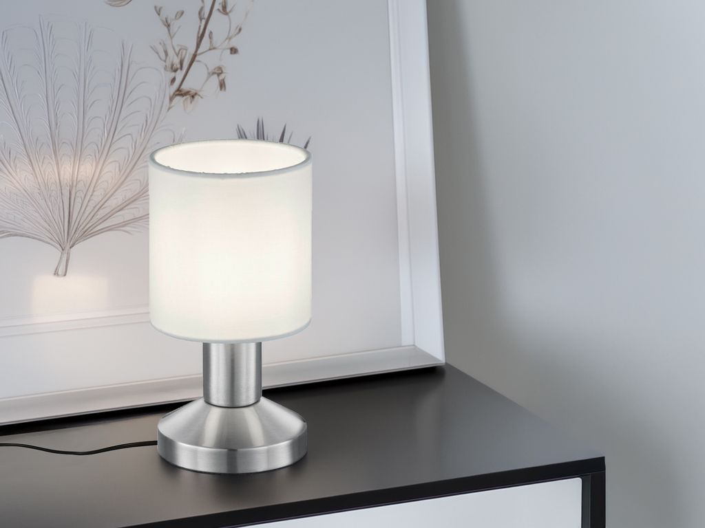A modern table lamp with a white fabric shade and a brushed silver base is placed on a dark surface next to a framed picture.
