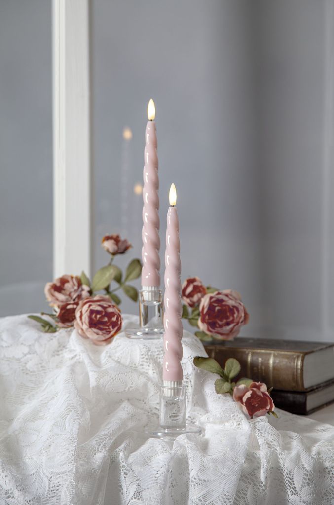 Two pink LED candles in glass holders are placed on a white lace tablecloth, surrounded by pink roses and two old books.