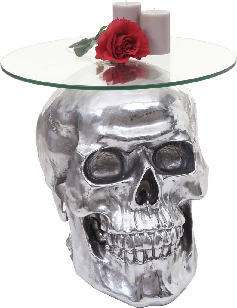 A silver skull-shaped side table with a glass top, a red rose, and two gray candles on it.
