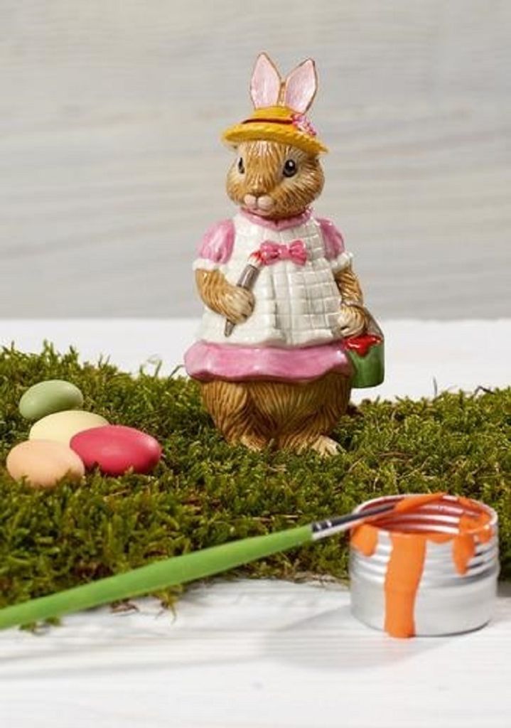 A ceramic Easter bunny figurine dressed in a hat and apron, holding a paintbrush and a bucket of paint, surrounded by moss and colorful Easter eggs.