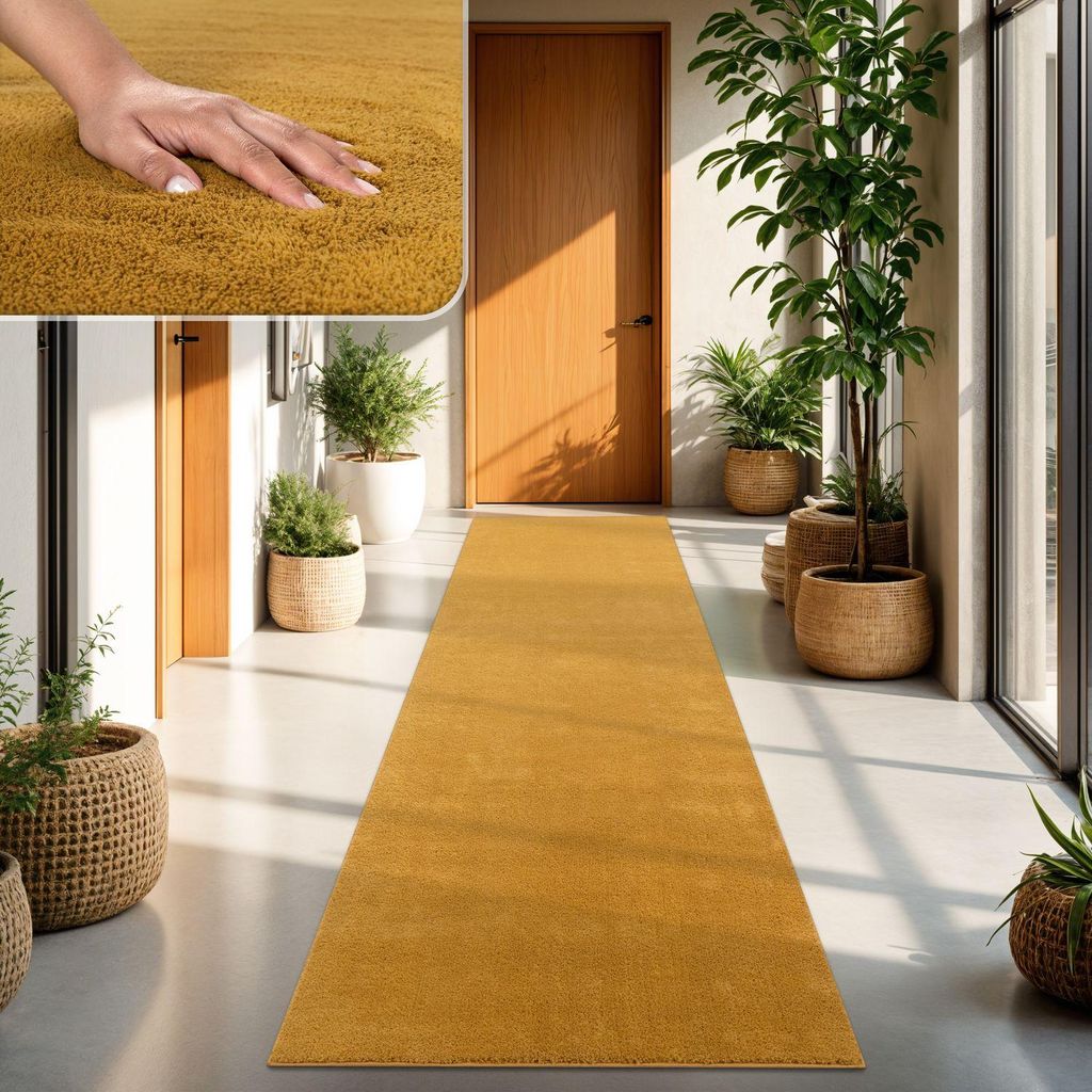 A hand touching a yellow rug in a hallway with plants and doors.