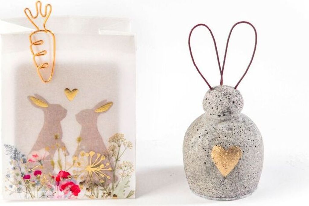 Two Easter-themed decorative items: a white paper bag with a carrot clip and bunny graphics, and a gray ceramic bunny bell with a gold heart.